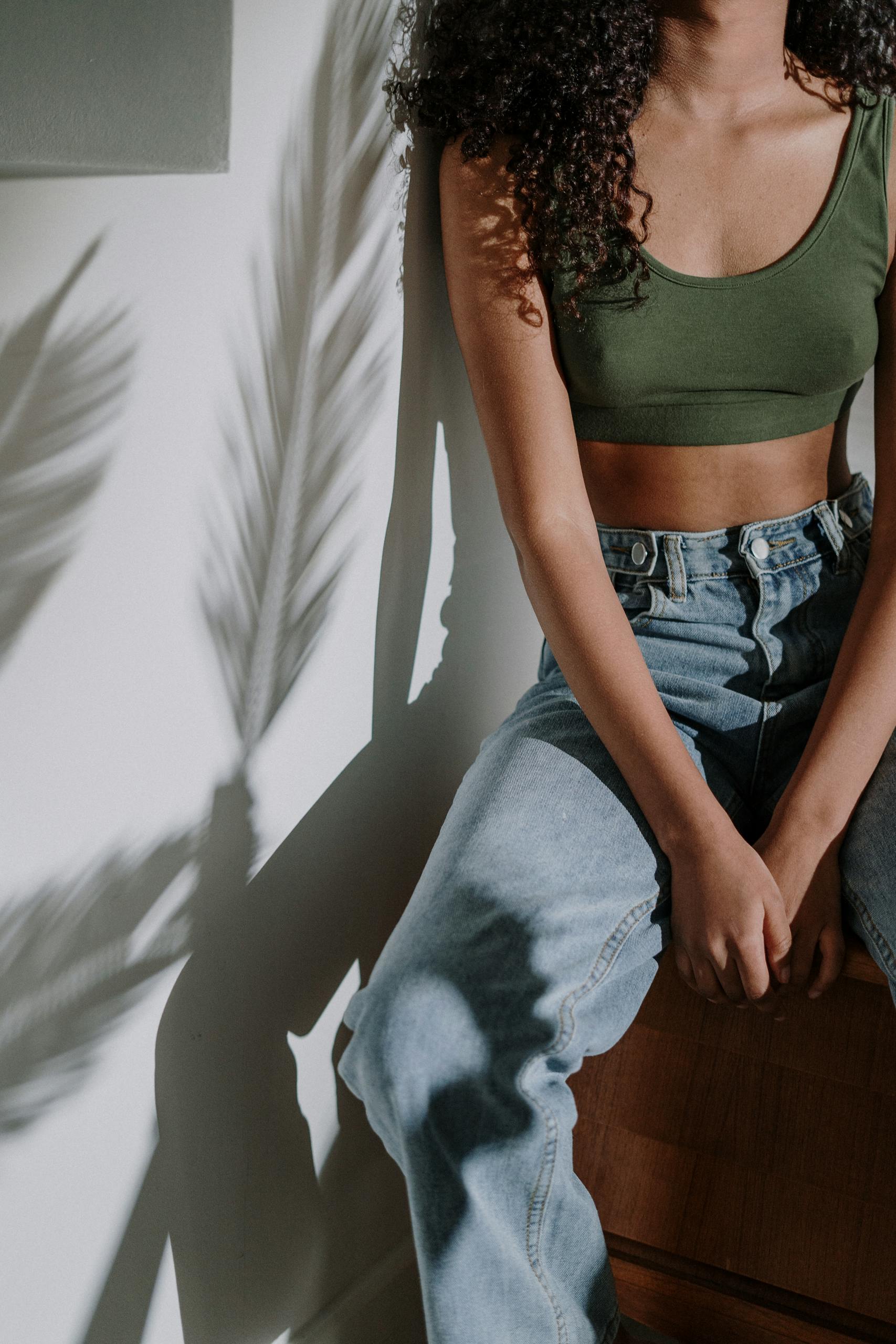 Home Woman sitting in sunlight wearing denim, with tropical plant shadows on the wall.