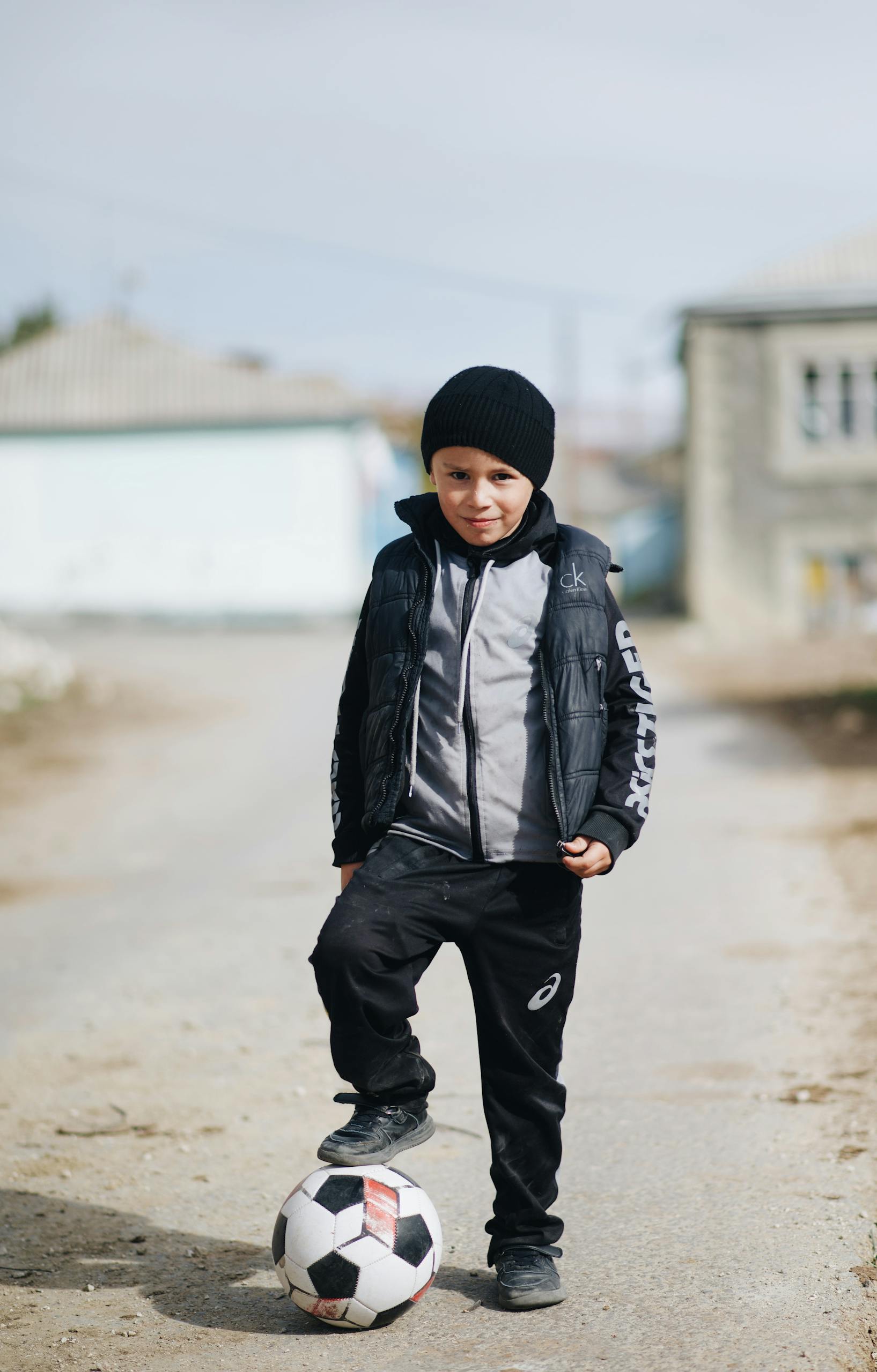 Home Young boy in warm clothing playing soccer outdoors on a street, showcasing childhood sports activities.